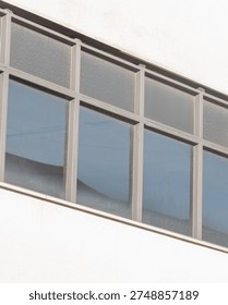Gray windows are arranged in a grid pattern on a white wall. The sunlight reflects off the glass, showing the blue sky above. The setting is urban and simple.
