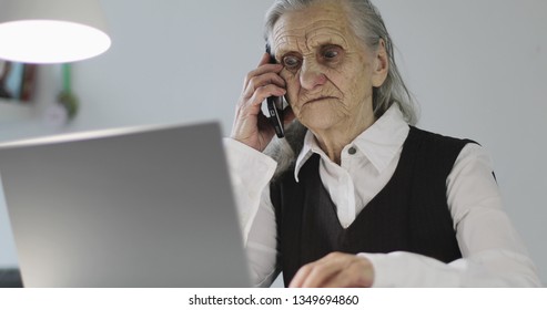 A Gray-haired Angry Old Woman Works At A Laptop And Talks On A Mobile Phone. Business Grandmother Sitting In An Office. COVID-19