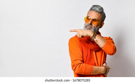 Gray-haired, bearded, old-aged man in orange sweater and scarf, sunglasses, bracelets. Pointing at something by his forefinger, looking aside, posing isolated on white. Close up, copy space