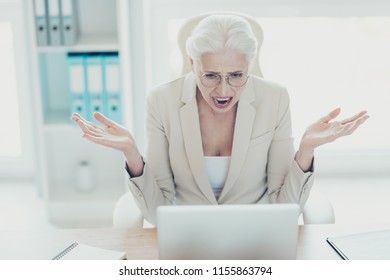 Gray-haired beautiful stylish mad crazy angry unhappy upset old lady wearing glasses, screaming, showing devastation, sitting in front of netbook at workplace workstation - Shutterstock ID 1155863794