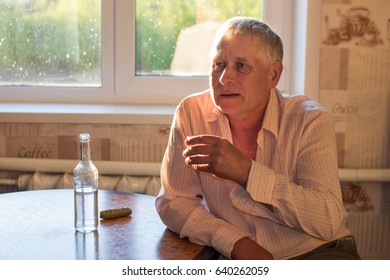A gray-haired gray man is sitting at the kitchen table, on which stands a bottle of alcohol, a pickled cucumber, and holds a glass of vodka in his hand. Social problems: alcoholism