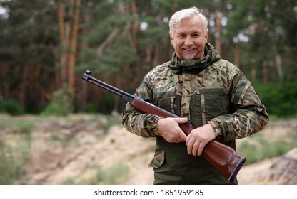 The gray-haired hunter stands with a gun in his hands in the wild forest and smiles at the camera. The hunter stands in the forest and holds a gun in his hands.