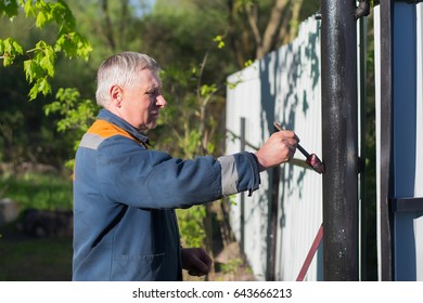 A Gray-haired Male Construction Worker Painting Black Metal Fence Pipe With A Brush