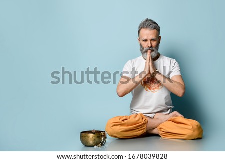 Gray-haired male, Hare Krishna follower, in traditional orange and white dhoti clothes. Praying, sitting on floor in lotus pose on blue background. Singing bowl, rosary nearby. Close up, copy space