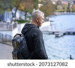 A gray-haired man with a backpack stands by a metal fence on the city embankment and looks at the water.