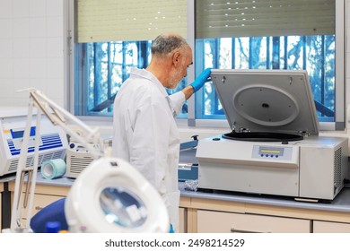 A gray-haired scientist, wearing a white lab coat and blue gloves, manipulates a centrifuge in a modern laboratory. The centrifuge is open, and the scientist appears to be placing or removing samples. - Powered by Shutterstock