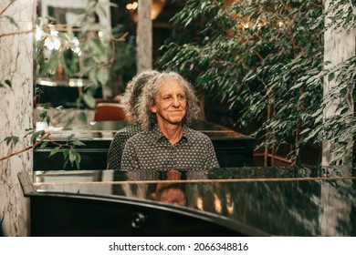  gray-haired senior man with curly long hair is sitting at piano and playing music, enjoying pleasant evening at hotel. 