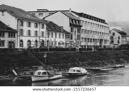 A grayscale shot of the boats on the river against the buildings on the shore 