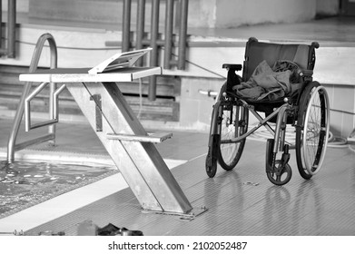 A Grayscale Shot Of A Wheelchair By A Diving Board At An Indoor Swimming Pool