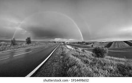 Grayscale. Spring rapeseed and small farmlands fields after rain evening view, cloudy pre sunset sky with rainbow and rural hills. Seasonal, weather, climate, eco, farming, countryside beauty concept.