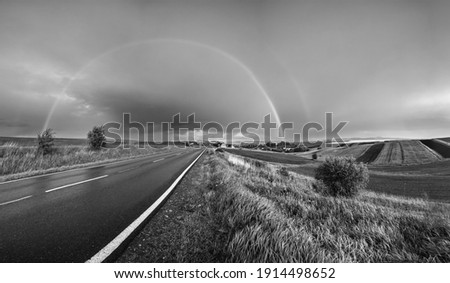 Grayscale. Spring rapeseed and small farmlands fields after rain evening view, cloudy pre sunset sky with rainbow and rural hills. Seasonal, weather, climate, eco, farming, countryside beauty concept.