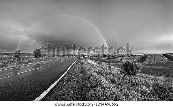 Grayscale. Spring rapeseed and small farmlands\
fields after rain evening view, cloudy pre sunset sky with rainbow\
and rural hills. Seasonal, weather, climate, eco, farming,\
countryside beauty\
concept.