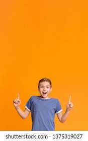 It's great. Amazed smiling teen boy pointing two fingers up at empty space, orange studio background