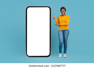 Great App. Beautiful African American Woman Pointing At Big Blank Mobile Phone, Young Black Female Demonstrating Smartphone With White Screen For Online Advertisement, Blue Background, Mockup