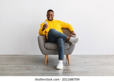 Great App. Contented Black Man Using Smartphone Advertising New Mobile Application, Texting Online Sitting In Armchair Near Gray Wall Indoors. Modern Mobile Communication, People And Gadgets - Powered by Shutterstock