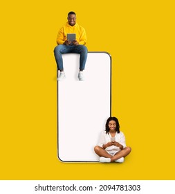 Great App. Excited Black Couple Sitting On Big Smartphone With Blank White Screen Using Gadgets, Cheerful Guy And Lady Chatting On Social Media, Yellow Orange Wall, Mock Up. Modern Communication
