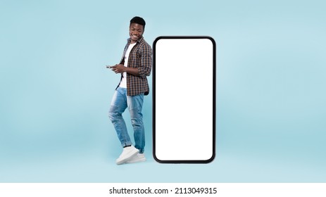 Great App. Excited Black Man Leaning On Big Smartphone With Blank White Screen Using His Cell Phone, Cheerful Guy Chatting On Social Media, Standing On Blue Background, Mock Up Image, Full Body Length