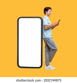 Great App. Excited Man Leaning On Big Smartphone With Blank White Screen And Using His Cell Phone, Cheerful Guy Chatting On Social Media, Standing On Yellow Background, Mock Up Image, Full Body Length