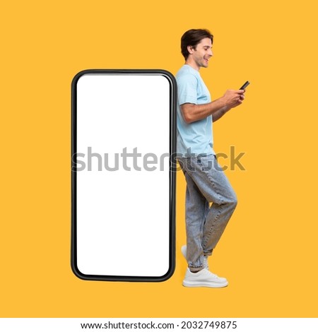 Great App. Excited Man Leaning On Big Smartphone With Blank White Screen And Using His Cell Phone, Cheerful Guy Chatting On Social Media, Standing On Yellow Background, Mock Up Image, Full Body Length