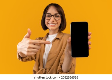Great App. Excited Woman Pointing At Smartphone Blank Screen, Demonstrating Free Space For Mobile App Or Website Design, Standing On Yellow Background, Mockup