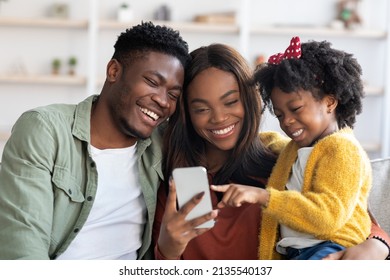 Great App. Happy Black Family With Little Daughter Using Smartphone At Home, Cheerful African American Mom, Dad And Female Child Looking At Mobile Phone Screen And Smiling, Closeup Shot