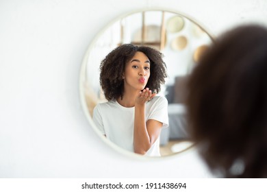 Great appearance, love yourself and perfect skin. Cheerful young african american lady makes air kiss and looks in reflection of round mirror in living room, on white wall, panorama, free space - Shutterstock ID 1911438694