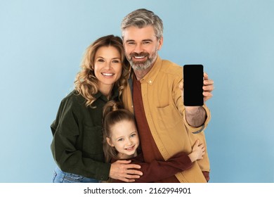 Great Application. Family Holding Mobile Phone Showing Blank Screen To Camera Recommending App Posing In Studio On Blue Background. Parents And Daughter Advertising Offer Via Smartphone. Mockup