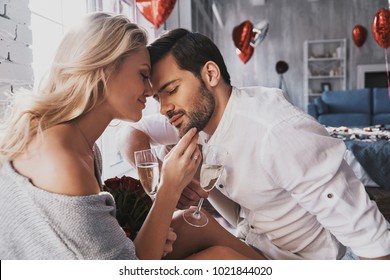 So great to be together! Beautiful young couple drinking champagne and smiling while sitting face to face in the bedroom - Shutterstock ID 1021844020