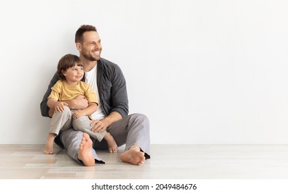 Great Family Ad. Happy Caucasian Father Carrying His Little Son, Sitting Together On Floor And Looking Aside At Empty Space, White Wall Background, Mockup For Text