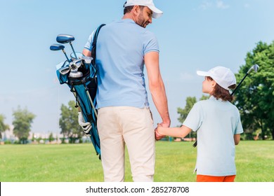 It was a great game! Rear view of young man and his son holding hands and looking at each other while walking on the golf course