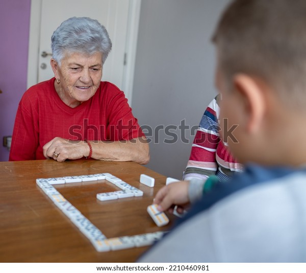 great grandson and\
great-grandmother are playing dominoes. the boy is making a move,\
the great-grandmother is watching his next move while there are\
dominoes on the table