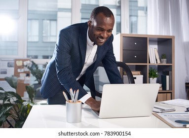 Great job. Positive satisfied young african businessman in suit is standing in office while leaning over table and typing on laptop with smile. Big window in background - Powered by Shutterstock