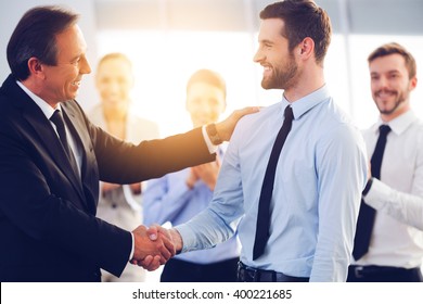 Great Job! Two Cheerful Business Men Shaking Hands While Their Colleagues Applauding And Smiling In The Background