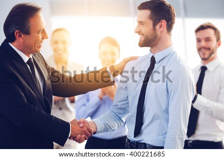 Great job! Two cheerful business men shaking hands while their colleagues applauding and smiling in the background