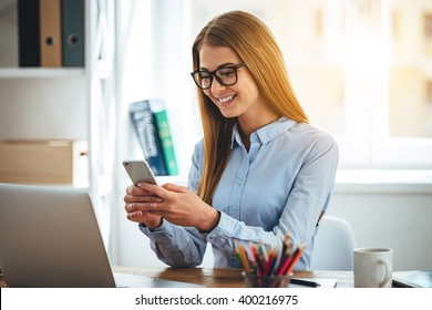 Great message! Cheerful young beautiful woman in glasses using her smartphone with smile while sitting at her working place - Powered by Shutterstock