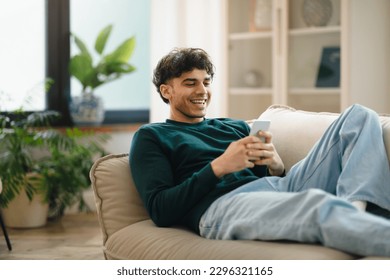 Great Mobile Offer. Cheerful Arabic Man Using Phone Lying On Couch Relaxing At Home On Weekend. Guy Using New Application On Smartphone, Browsing Internet And Texting Online. Gadgets Lifestyle - Powered by Shutterstock