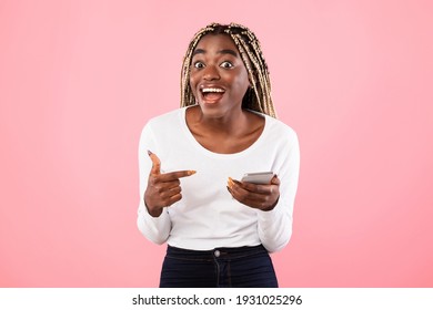 Great Mobile Offer. Excited Black Lady Pointing At Smartphone In Her Hands, Emotionally Reacting To New App, Overjoyed Millennial African American Woman Standing Isolated Over Pink Studio Background