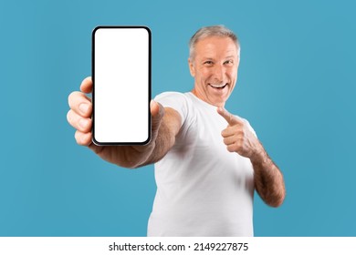 Great Mobile Offer. Excited Senior Man Holding Showing Big Cell Phone Close To Camera Pointing Finger At Smartphone In His Hand, Cheerful Adult Male Standing Isolated Over Blue Studio Background Wall