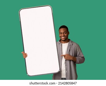 Great mobile offer. Happy black man pointing at huge empty smartphone in his hand, advertising new app or website, showing mockup for your design, standing over green background
