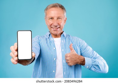 Great Mobile Offer. Happy Senior Man Holding Smartphone With Blank White Cell Screen And Gesturing Thumb Up Sign, Cheerful Adult Male Recommending New App Or Website, Blue Background, Mock Up Template