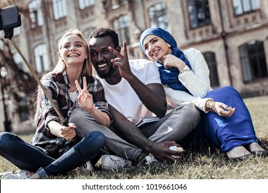 Great mood. Cheerful positive emotional student showing V sign and looking into the smartphone camera while taking a selfie together