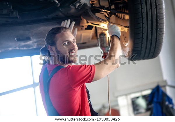 Great mood, work. Joyful\
smiling bearded man in overalls repairing car in auto repair shop\
in afternoon