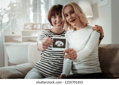 Great News. Beaming Same Sex Couple Showing Ultrasound Photo Of Their Future Baby Holding It With Both Hands