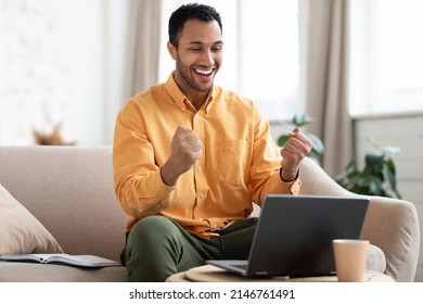 Great News Concept. Joyful guy using pc sitting on couch in living room looking at screen, male raising hands with excitement, ecstatic enthusiastic man shaking clenched fists celebrating success