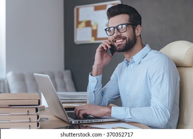 Great News! Handsome Cheerful Young Man Wearing Glasses Talking On Mobile Phone And Looking Away With Smile While Sitting At His Working Place