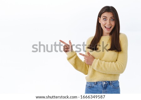 Great news hey take look. Excited happy and surprised beautiful young woman in yellow sweater turn yout attention at banner, pointing fingers left and scream joyful, stand white background