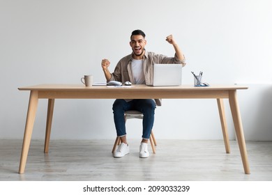 Great News Online. Portrait of joyful excited Arabic man raising hands with excitement, celebrating success shaking clenched fists sitting at table at home office using laptop looking at camera