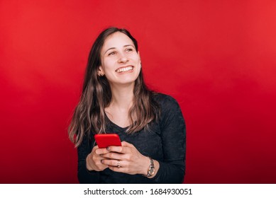 Great news. Smiling young woman looks up while she is holding her smart phone on red background