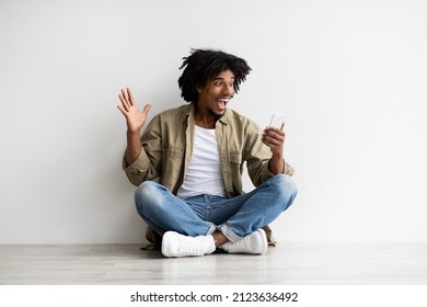 Great News. Surprised Young Black Guy Looking At Smartphone Screen With Excitement While Sitting On Floor Indoors, Emotional African American Man Raising Fist With Excitement, Copy Space