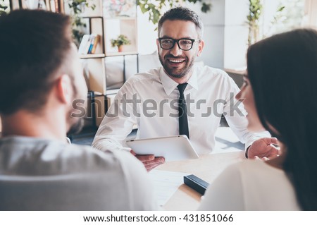 Great news for you! Cheerful mature man in shirt and tie holding digital tablet and gesturing while young couple sitting in front of him at the desk 